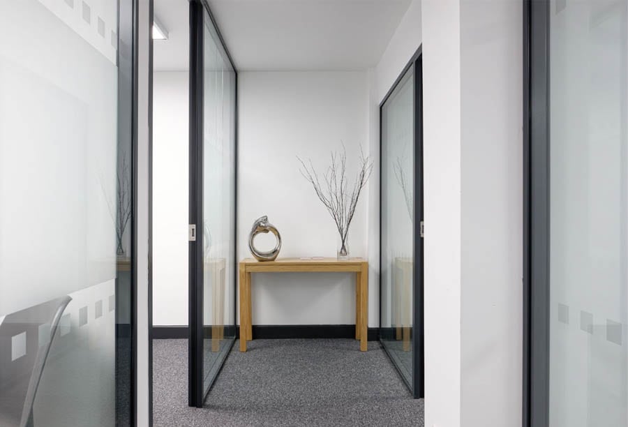 Glass partition walls | Corridor in office building with a wooden table at one end, glass walls on either side. 